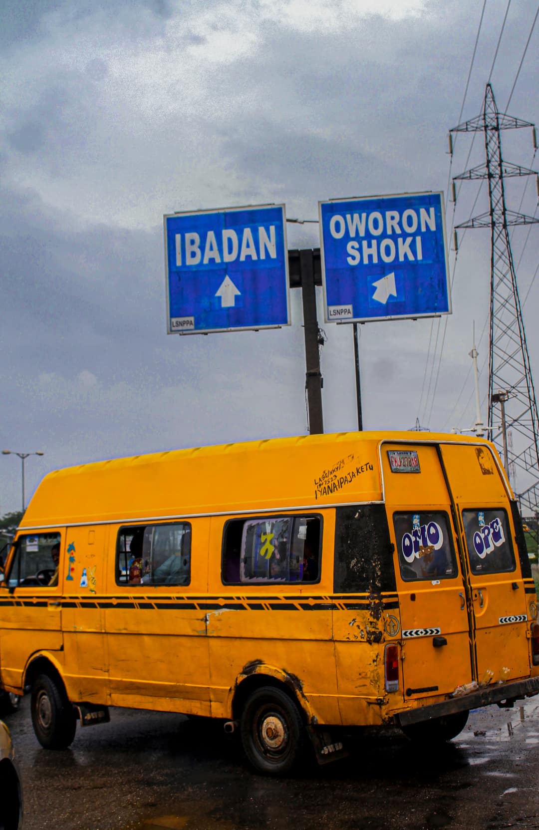 A yellow danfo passing under blue road signs for Ibadan and Oworonshoki